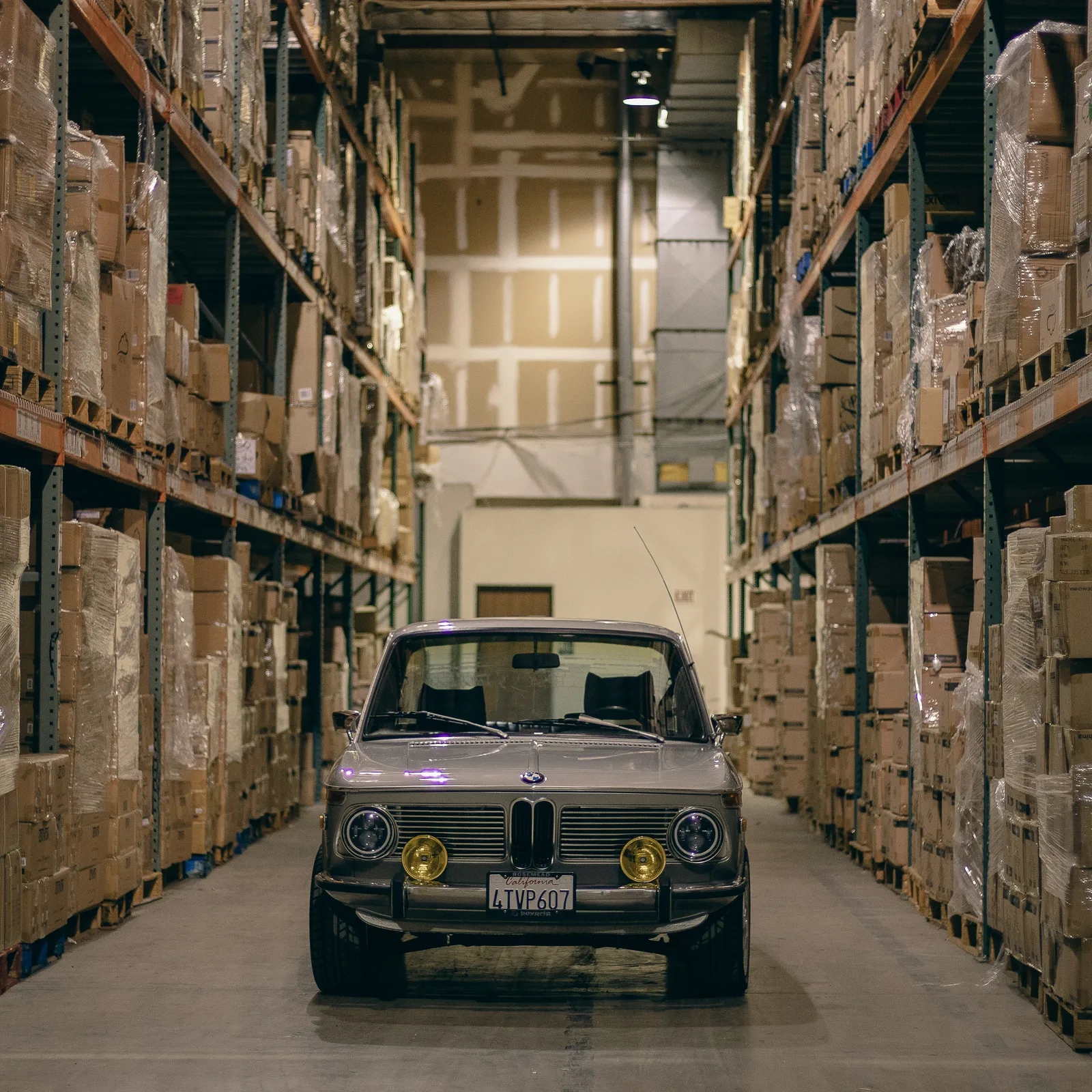 Classic BMW 2002 parked in a Detroit auto parts warehouse aisle between towering pallet racks of inventory